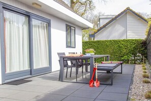 A modern house with a patio area featuring a table and chairs, a red shovel, and a hedge. - De Golbreker Type 1AC (Zoutelande)