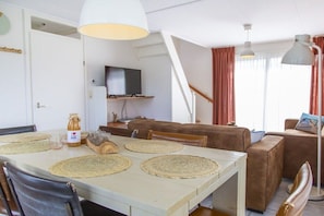 A dining area with a table set with placemats, a bottle of olive oil, and a television in the background. - De Golbreker Type 1AC (Zoutelande)