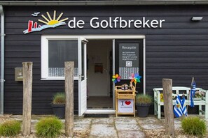 A black building with a sign reading 'de Golfbreker' and a reception area with a sign that reads 'de Golfbreker Wim Koppejan Margriet Louise de Golfbreker.nl'. There is a wooden bench with flags and a small table with a sign that says 'I love ZOUTELANDE'. - The Golf Breaker - Type 1 (Zoutelande)