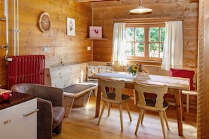 A wooden cabin interior with a dining table, chairs, a bench, a refrigerator, a window with curtains, and a wall-mounted clock. - Pifrailerhof "Alte Mühle" (Schnals - Südtirol)