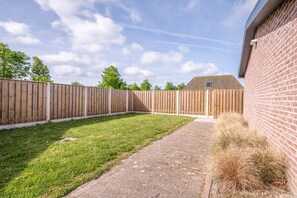 A fenced backyard with a paved path, a grassy area, and a brick building. - Holiday home with 1 bedroom (Oostkapelle)