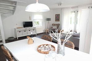 A modern living room with a white sofa, a dining table with a basket of snacks, and a television on a white cabinet. - Holiday home "Zee Gras" (Kamperland)