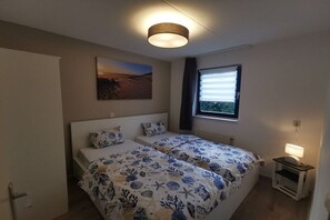 A bedroom with two beds, a painting on the wall, a window with blinds, and a bedside lamp. - Holiday home with 1 bedroom (Julianadorp)