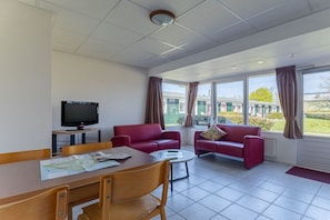 Ein modernes Hotelzimmer mit Fernseher, einem Esstisch mit Stühlen, zwei roten Sofas und großen, mit Vorhängen versehenen Fenstern.