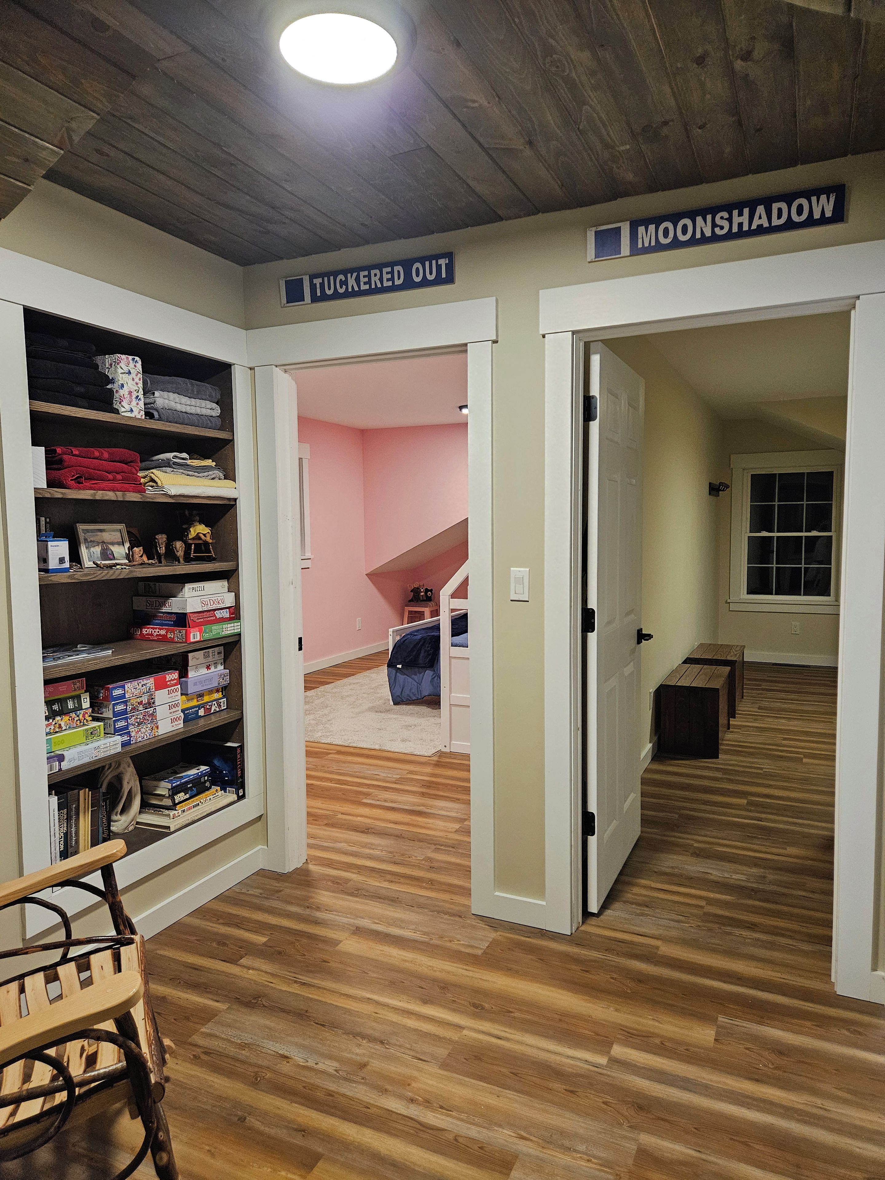 A hallway with a wooden floor, a bookshelf filled with board games, and a sign indicating 'MOONSHADOW' and 'TUCKERED OUT' areas.