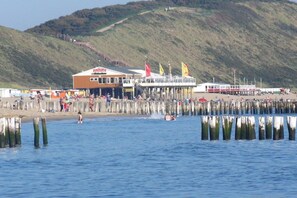 A coastal building with a pier extending into the water, surrounded by a sandy beach and hills in the background. - Holiday apartment in Zoutelande with Wi-Fi (Zoutelande)