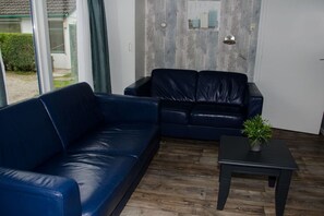 Quarto com sofá de couro azul, uma mesinha com uma planta e vista para uma casa através de uma porta de vidro.