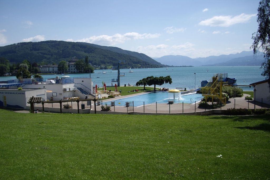 Een zwembad met uitzicht op een meer en bergen, omringd door grasvelden en gebouwen.