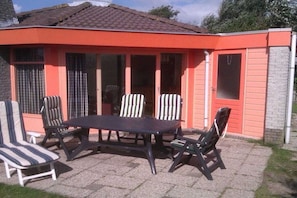 Une terrasse extérieure avec une table et des chaises, un espace couvert avec des fenêtres et un mur extérieur rouge.