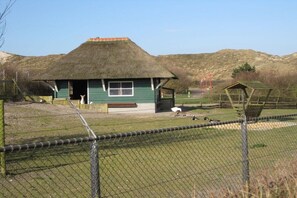 Een gebouw met rieten dak en een groene buitenkant, een omheind verblijf met vogels en een glooiend landschap op de achtergrond.