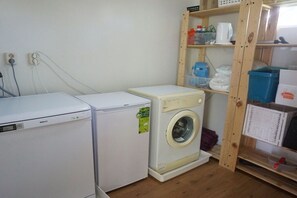 A laundry area with two washing machines, a wooden shelf with various items, and a power outlet on the wall. - Merel, De Keizerskroon 93 (Julianadorp aan Zee)