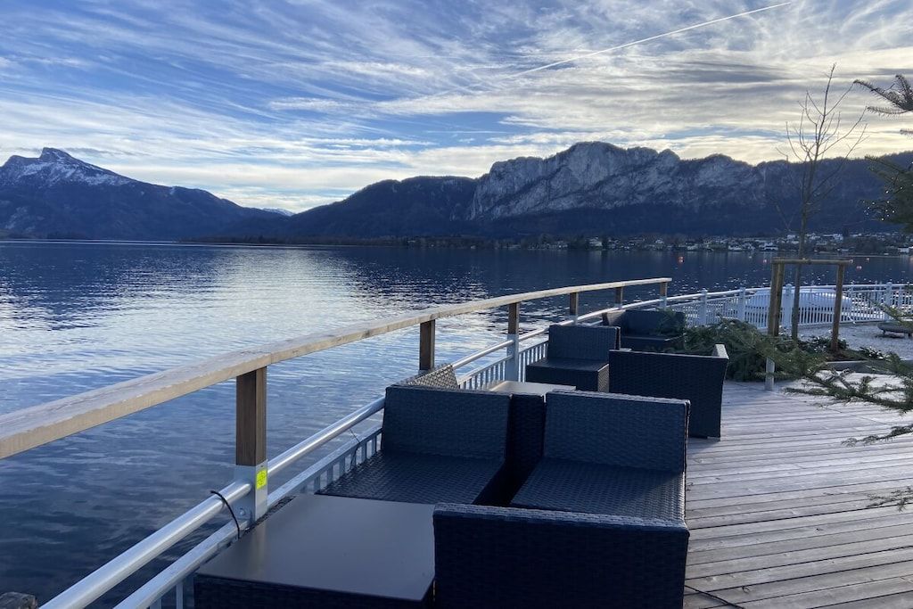 A deck with outdoor seating overlooking a calm lake and mountains.