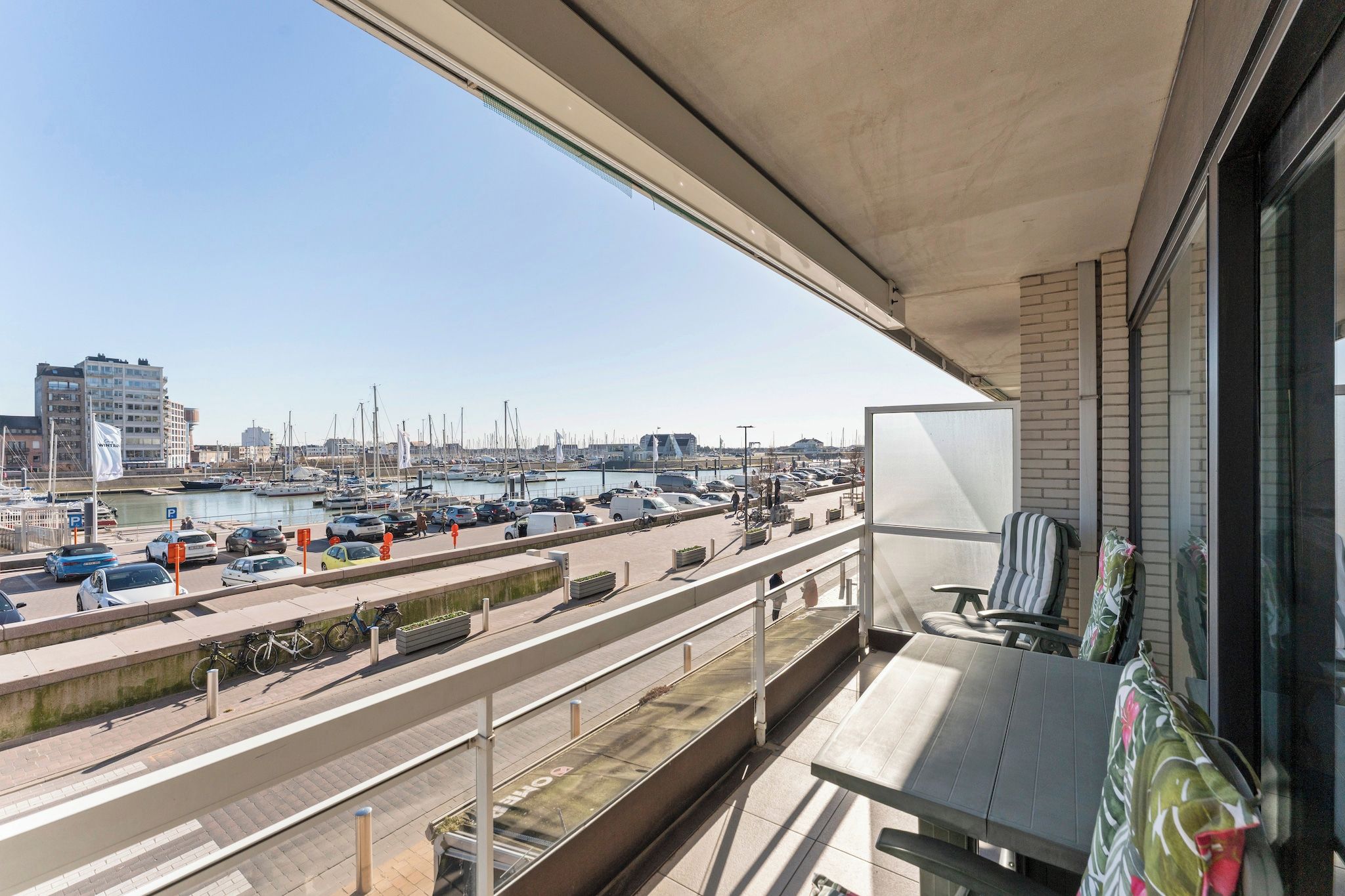 Un balcon donnant sur une marina, une route où circulent des véhicules et un bâtiment en arrière-plan.