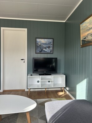 Ein Zimmer mit einer weißen Tür, einem Flachbildfernseher auf einem weißen Schrank, einem Gemälde über dem Fernseher und einem kleinen Tisch mit Kissen.