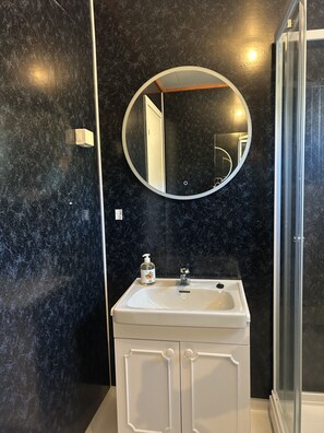 Ein Badezimmer mit einem weißen Waschbecken, einem runden Spiegel und einer Duschkabine aus Glas.