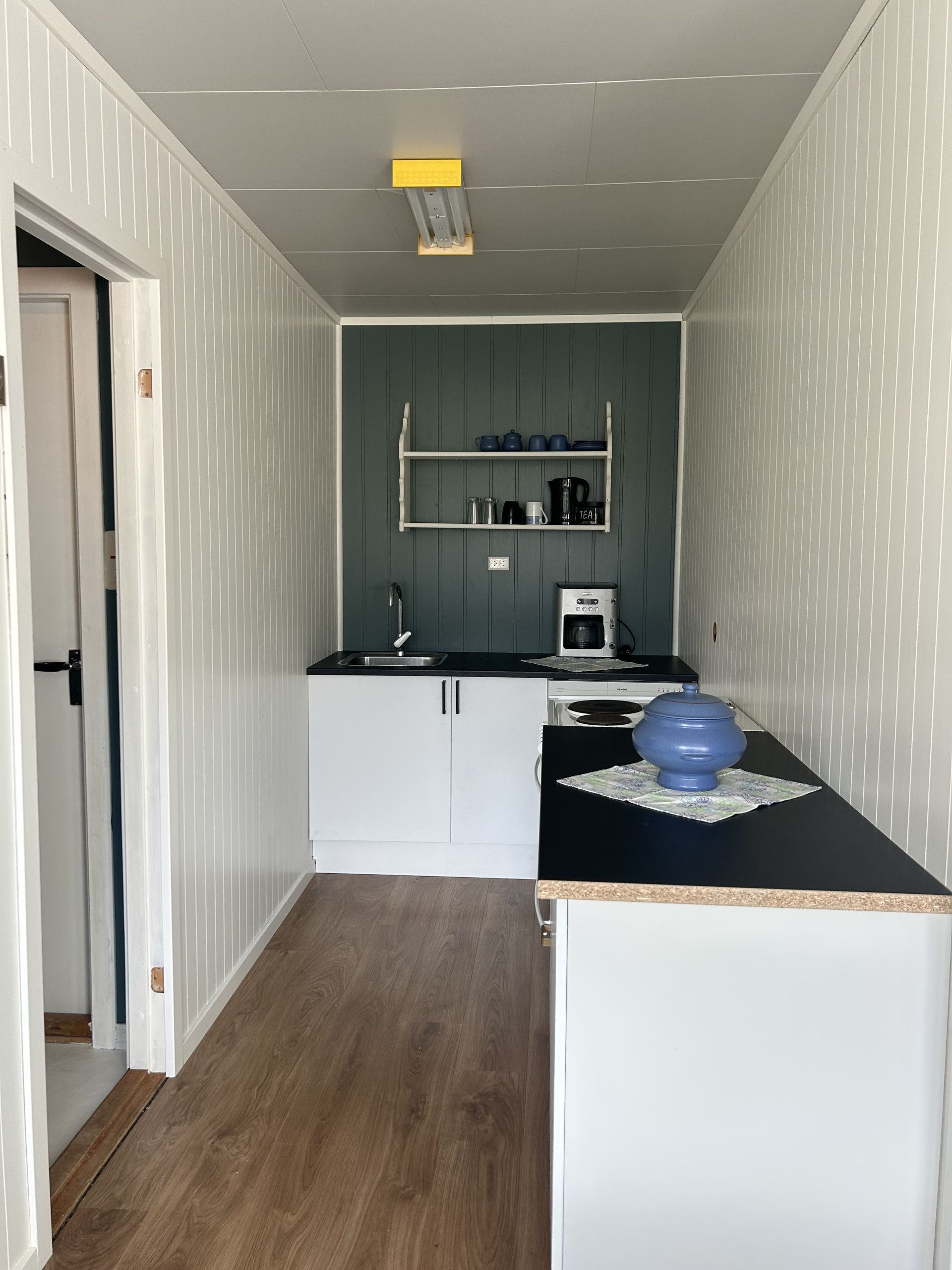 A compact kitchen with white cabinets, a black countertop, and a small sink. There is a shelf above the countertop holding kitchenware.