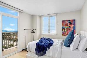 Een slaapkamer met een bed, een raam met zonwering, een kleurrijk abstract schilderij en uitzicht op een stadsgezicht vanaf een balkon.