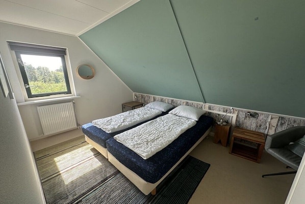 Een slaapkamer met een schuine kap, een raam, een bed met twee kussens, een nachtkastje en een stoel.