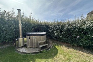 Een kuip met warm water, met een houten buitenkant en een metalen verwarming, omringd door een houten terras en een haag.