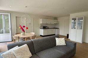 A modern living room with a grey sofa, a dining table with chairs, a kitchen area with white cabinets, and a glass cabinet. - Holiday apartment with 3 bedrooms (Callantsoog)