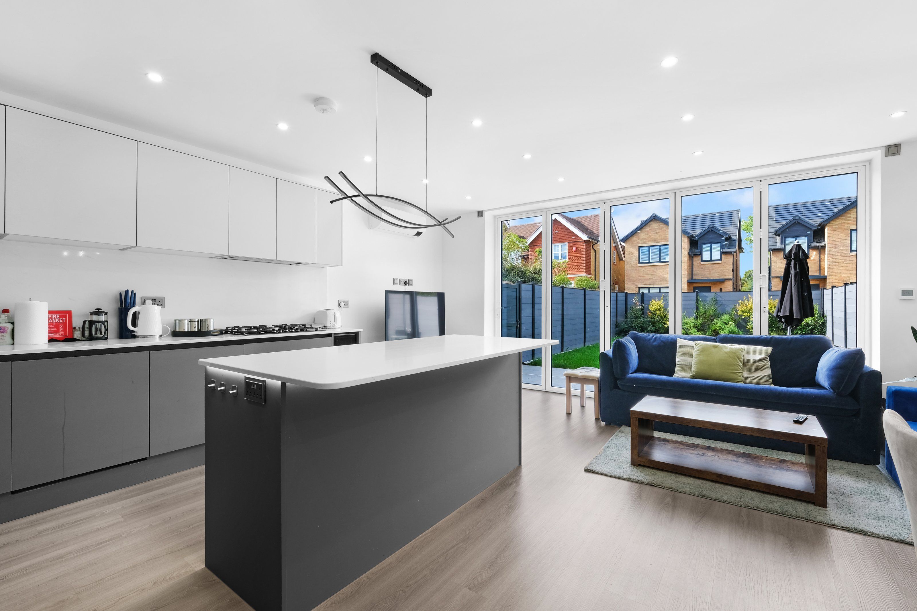 A modern kitchen with a central island, a dining area with a blue sofa, and a view of a residential neighborhood through large sliding doors.