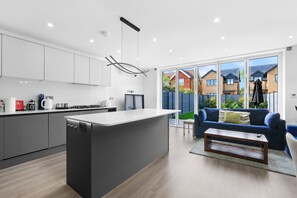 Una cocina moderna con una isla central, un comedor con un sofá azul y vistas a un barrio residencial a través de amplios ventanales correderas.