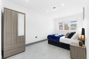 A bedroom with a bed, bedside table, lamp, and a wardrobe. - Luxaura Homes 6 Bed Detached House (Maidenhead)