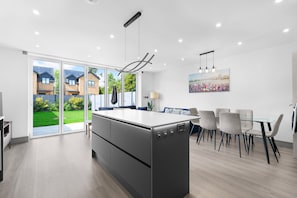 A modern kitchen with a large island, a dining area with a glass table, and a view of a residential area through glass doors. - Luxaura Homes 6 Bed Detached House (Maidenhead)