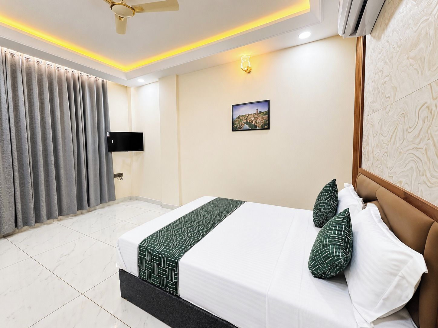 A hotel room with a bed, curtains, a wall-mounted TV, a picture, and a ceiling fan.