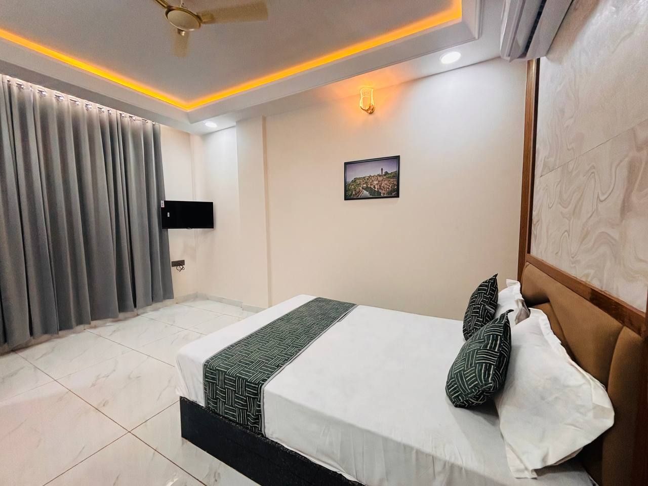 A hotel room with a bed, curtains, a flat-screen TV, a wall-mounted light, and a framed picture.