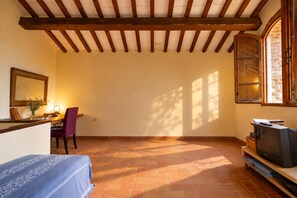 Habitación con piso de baldosas, techo de madera, televisor sobre un mueble de madera, una silla, una mesa con lámpara y una ventana con vistas.