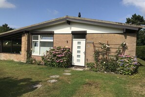 A single-story building with a white door and a garage door, surrounded by a well-maintained lawn and flowering plants. - "Holland House" at De Zandloper (Julianadorp)