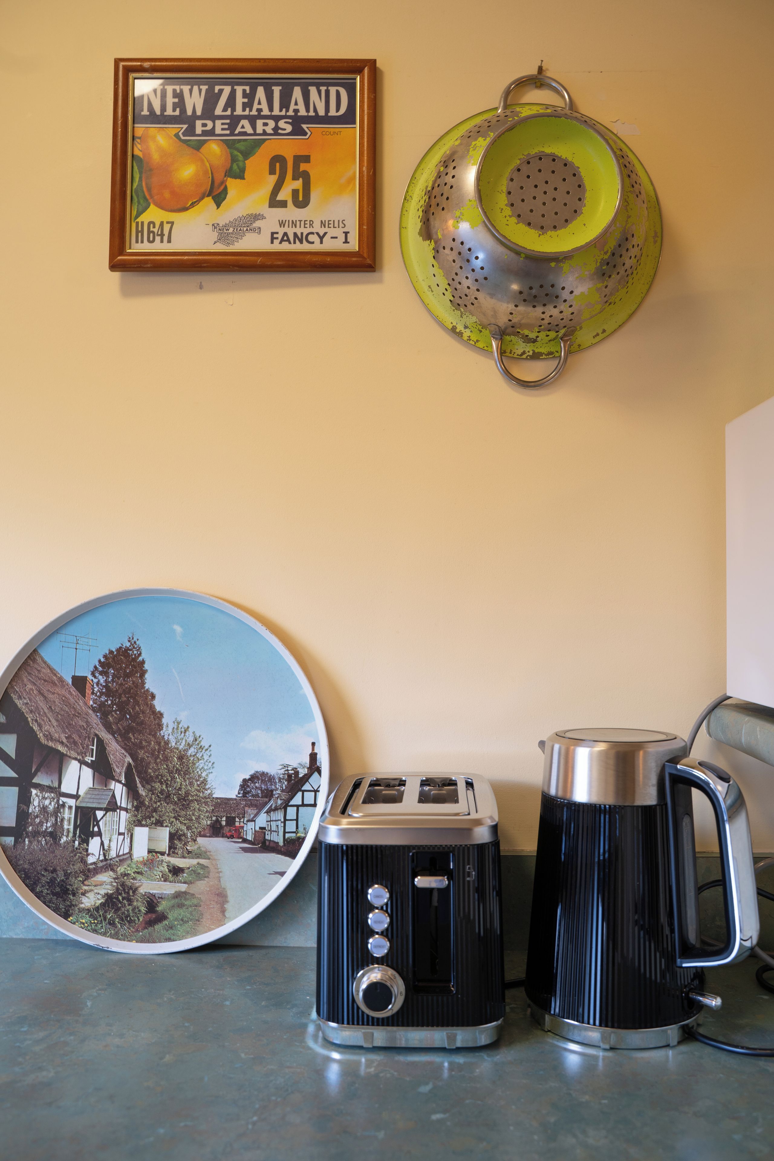 A kitchen counter with a vintage toaster, a kettle, a colander, and a framed picture of New Zealand pears.