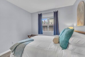 A bedroom with a bed, a window with curtains, a view of buildings outside, and a wall-mounted artwork. - Seagulls Nest | Steps 2 Lake! Massage Chair! (Sheboygan)