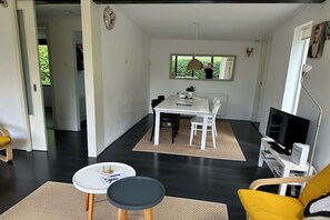 A modern living room with a dining area, a sofa, a TV, and a window with a view of greenery. - Holiday home with 3 bedrooms (Oostkapelle)