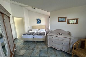 Een slaapkamer met een tweepersoonsbed, een dressoir en een stoel. Er is een deur die naar een andere kamer leidt.