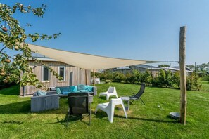 A backyard with a wooden house, outdoor furniture, and a large shade sail. - Duinpark Westerschouwen - Chalet 1 (Burgh-Haamstede)