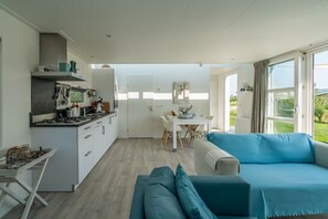 A modern kitchen with white cabinetry, a dining area with a white table and chairs, and a blue sofa in a bright room with large windows. - Duinpark Westerschouwen - Chalet 1 (Burgh-Haamstede)