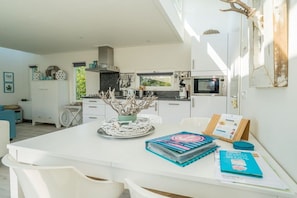 Une cuisine moderne avec des meubles blancs, un coin repas avec des chaises blanches et une vue sur l’extérieur.