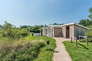 A building with a gravel path leading to a wooden structure, surrounded by greenery. - Duinpark Westerschouwen - Chalet 1 (Burgh-Haamstede)
