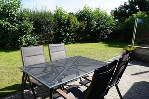 An outdoor patio with a metal table and chairs, surrounded by a grassy area and dense shrubbery. - Holiday home with 3 bedrooms (Julianadorp aan Zee)