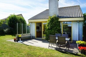 Una casa con techo de tejas, chimenea, puerta de entrada de vidrio, un patio con mesa y sillas, y un césped bien cuidado.