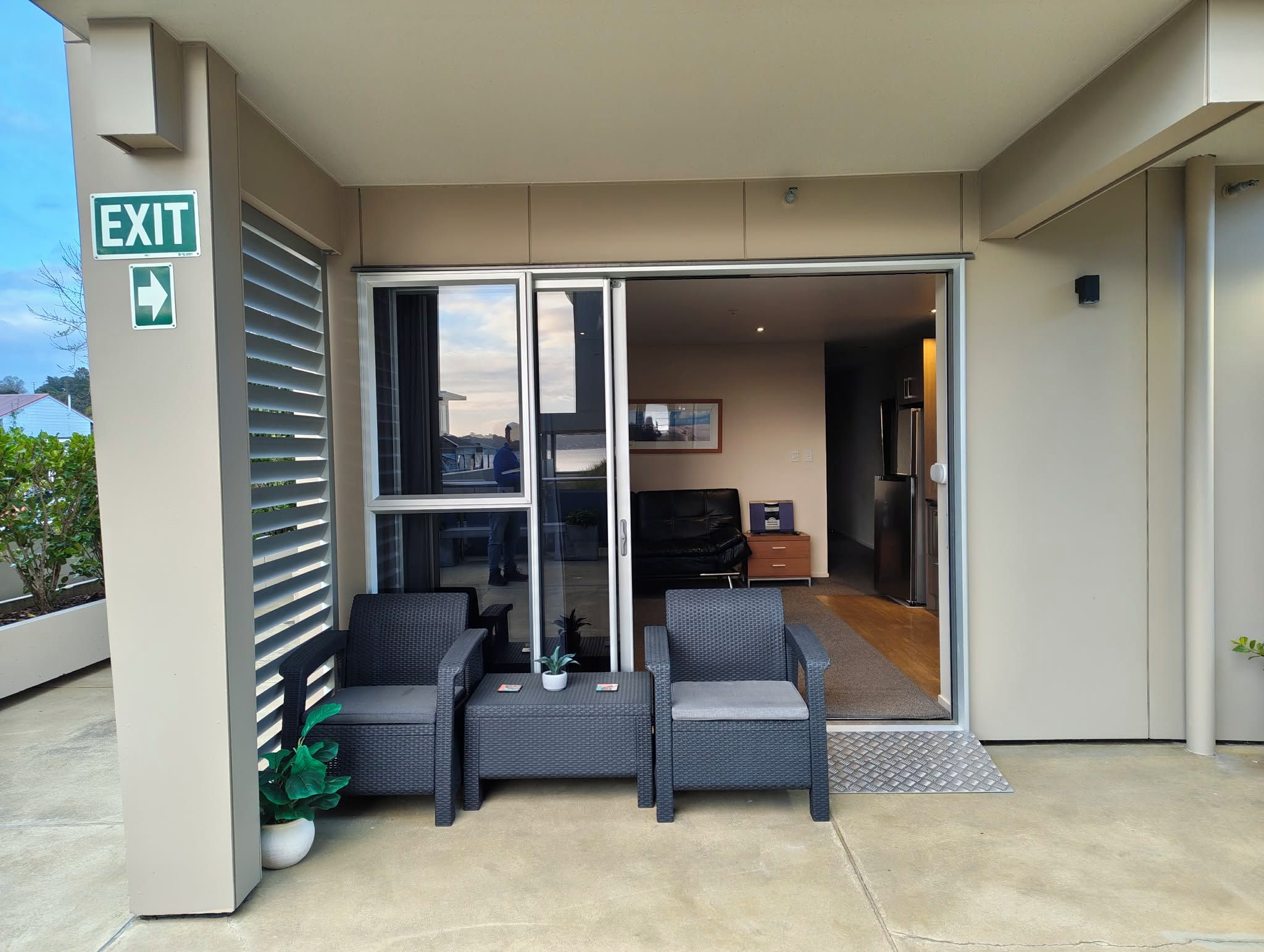 A hotel entrance with an exit sign, a glass door leading to a seating area with two chairs and a table, and a small potted plant.