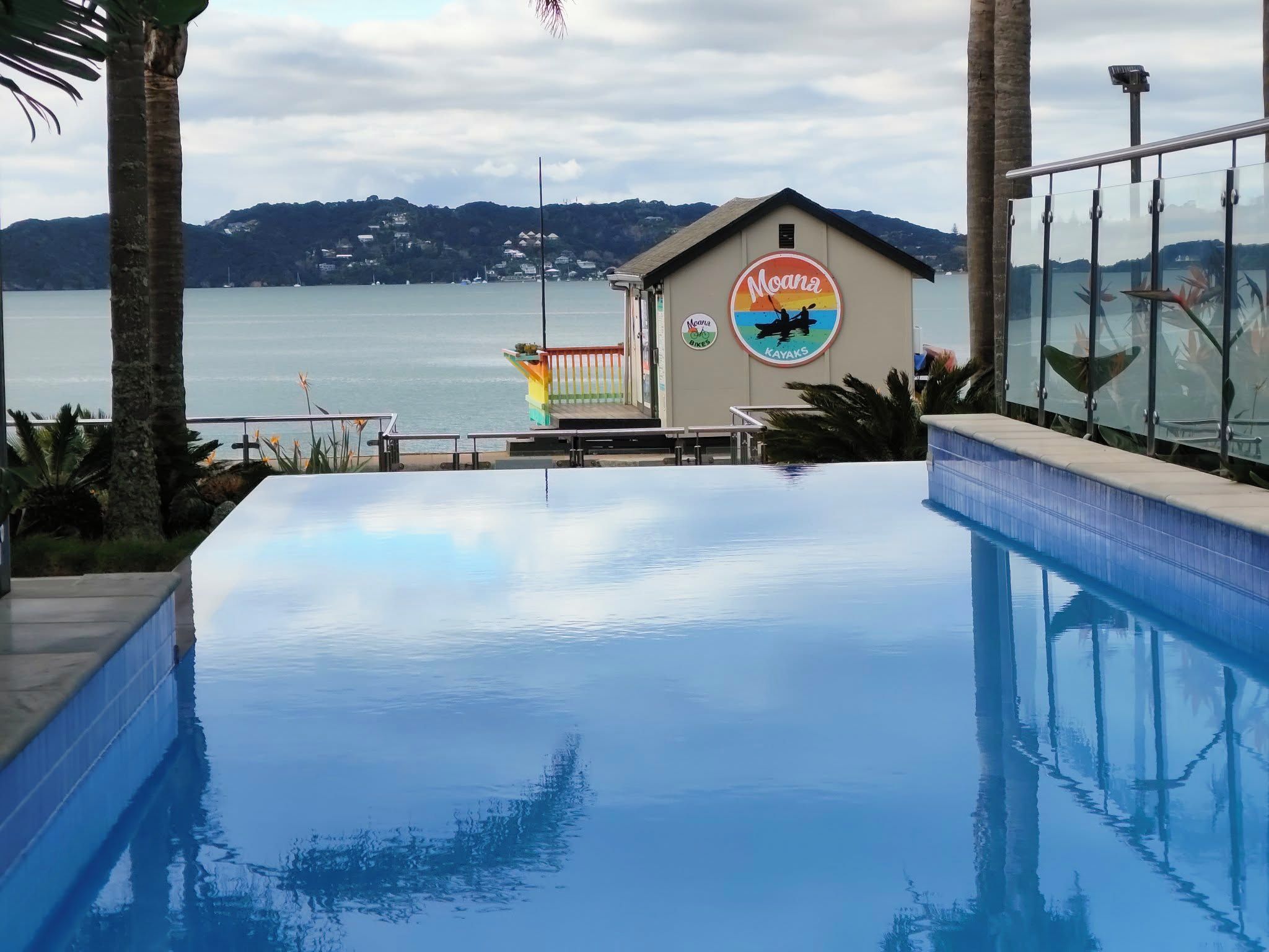 A swimming pool with a view of the sea and mountains, a building with a sign for 'Moana Bikes & Kayaks', and a glass railing.