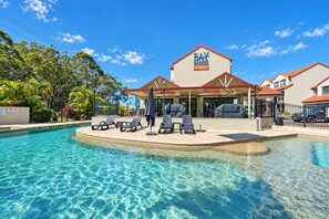 A pool area with lounge chairs and umbrellas, a building with a sign reading 'Bay Breeze Loft Apartments', and a clear blue sky. - Breeze at Fly Point Apartment 39 (Nelson Bay)