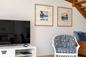 Ein Wohnzimmer mit einem Flachbildfernseher auf einem weißen Standfuß, zwei gerahmten Strandbildern, einem gemusterten Sessel und einer Holztreppe.