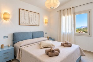 Ein Schlafzimmer mit Bett, Nachttischen, einer Wanddekoration, einem Fenster mit Vorhängen und Blick auf die Landschaft.