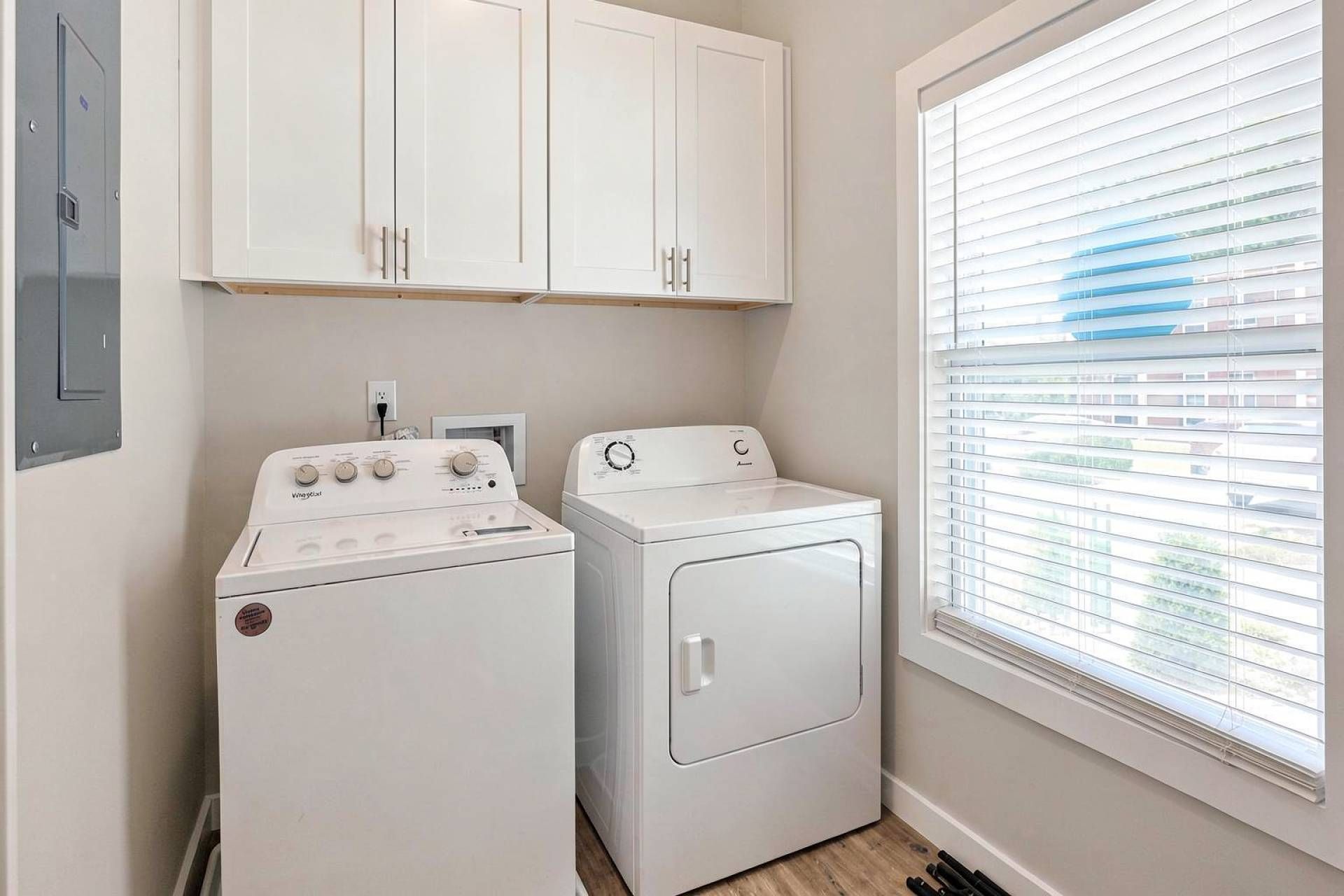 A laundry room with a washer and dryer, cabinets above, and a window with blinds.