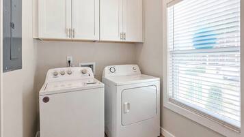 A laundry room with a washer and dryer, cabinets above, and a window with blinds.