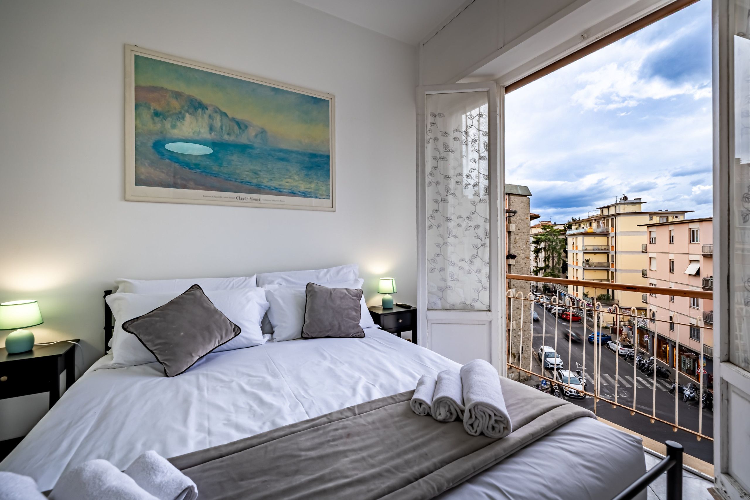 Quarto com cama, almofadas, manta e uma pintura de uma paisagem costeira.